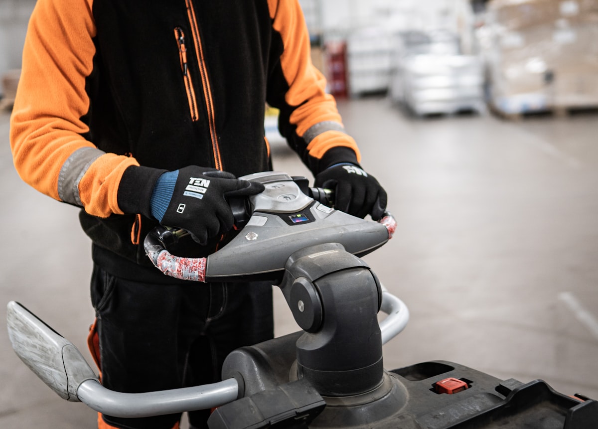 Technician in a safety vest steering a walk-behind floor scrubber across a warehouse floor
